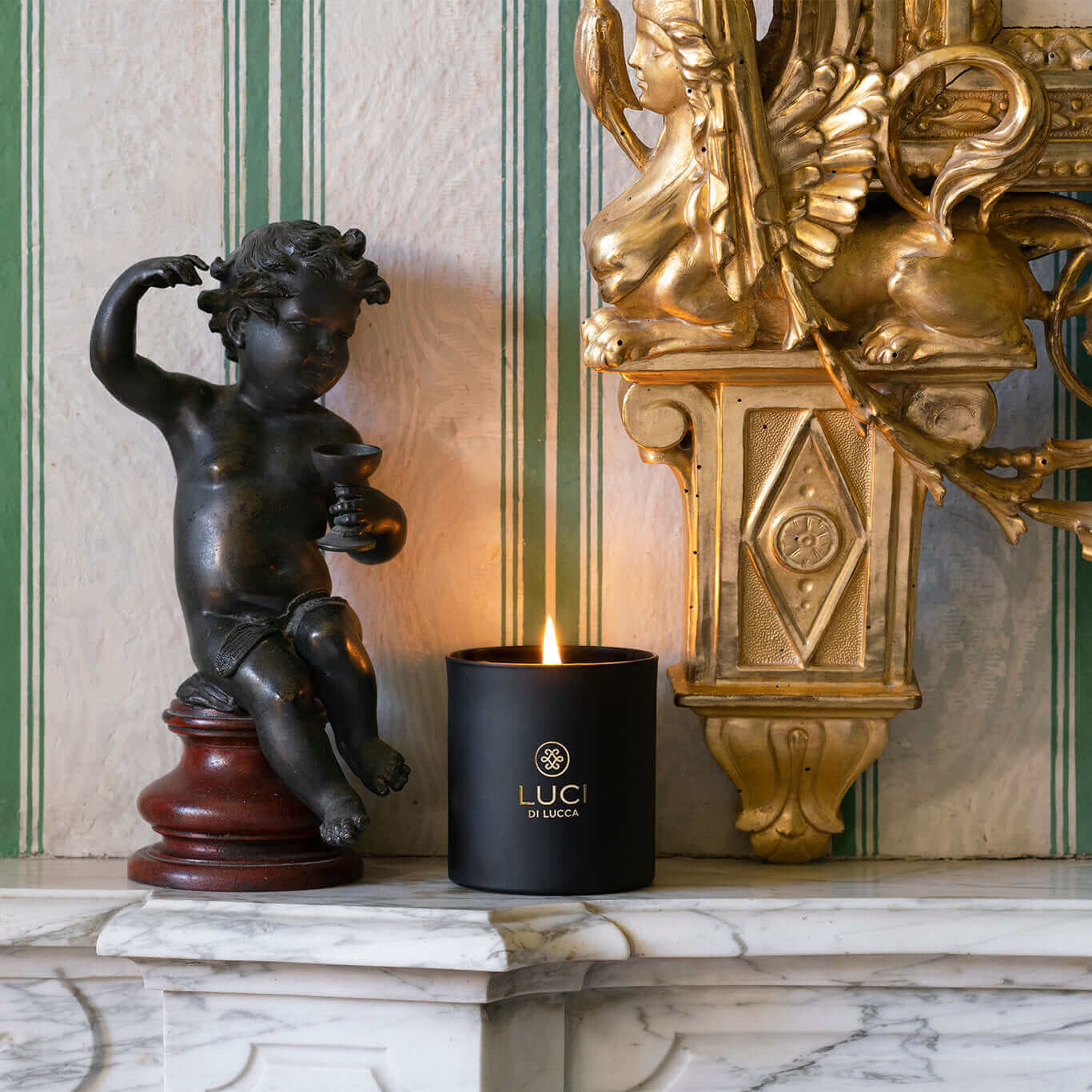 Luxury scented candle in black matte with gold details, flickering on a marble mantle in a villa setting, accompanied by a black cupid sculpture and gold architectural detail.