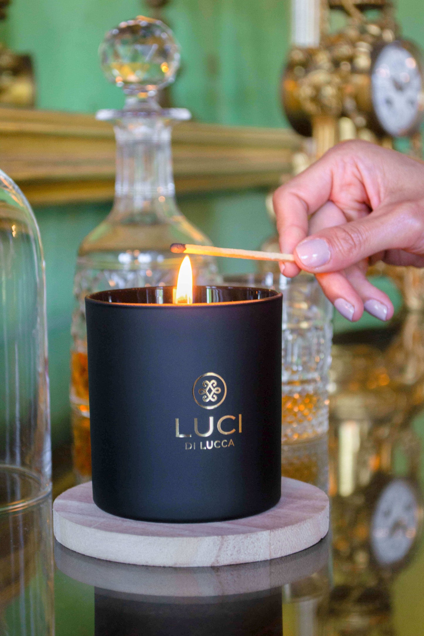 Hand lighting luxury scented candle with antique glassware, including a decanter, in the background.