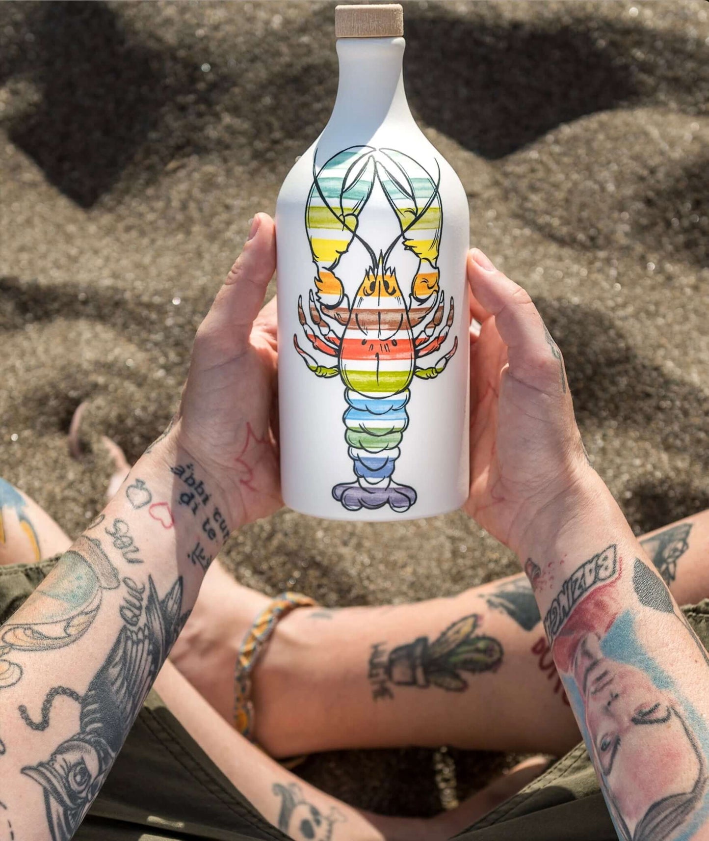 Rainbow lobster design olive oil bottle held on sandy beach, showcasing vibrant Mediterranean-inspired craftsmanship, with tattooed hand adding a unique touch.